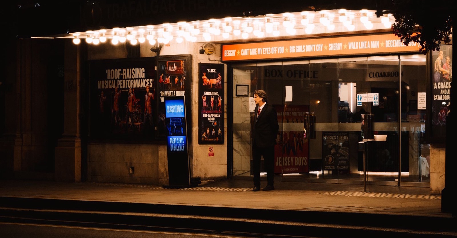 A lit box office at a theatre entrance at night, with one person waiting outside on the pavement