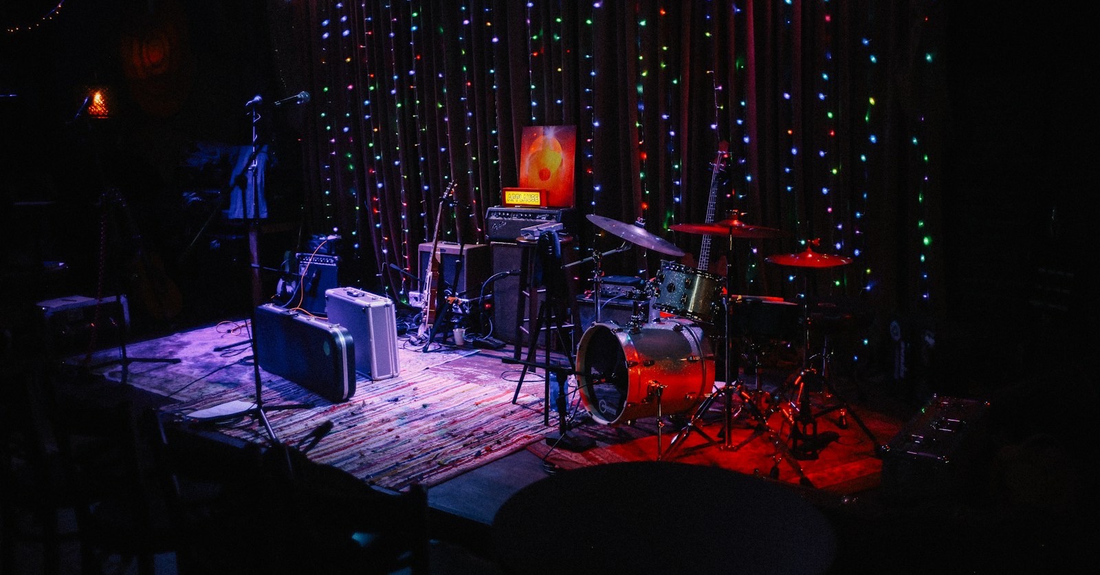 An empty club stage with a drum kit, amplifiers and guitar cases in front of a curtain of coloured fairy lights